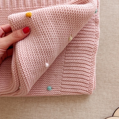 Pano de malha rosa com pompons coloridos segurado por mão com unhas vermelhas sobre tecido bege