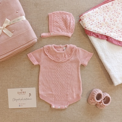 Conjunto de roupa de bebé rosa com body, gorro, sapatinhos e mantas sobre fundo bege