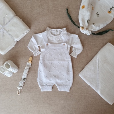 Conjunto completo de roupa e acessórios de bebé em branco e bege com detalhes florais num fundo bege.