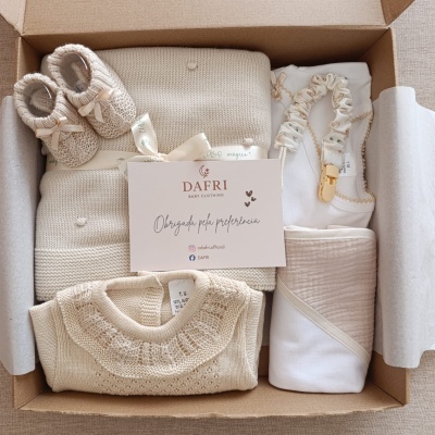 Conjunto de roupa e acessórios para bebé em tons bege e branco numa caixa de presente aberta.