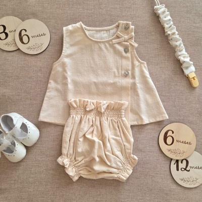 Conjunto de roupa de bebé bege com blusa, calções e sapatinhos brancos em fundo neutro com marcadores de idade em madeira
