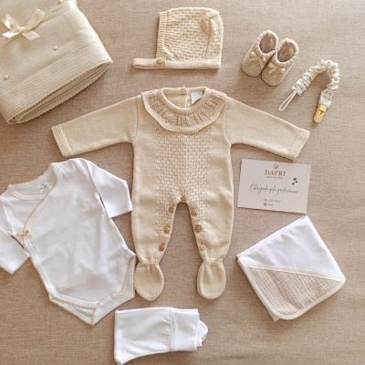 Conjunto de roupa para bebé em tons bege e branco com diferentes peças distribuídas sobre superfície neutra.