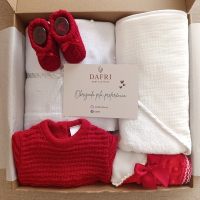 Conjunto de roupa de bebé com sapatinhos vermelhos, camisola vermelha e manta branca numa caixa de cartão com cartão da marca DAFRI