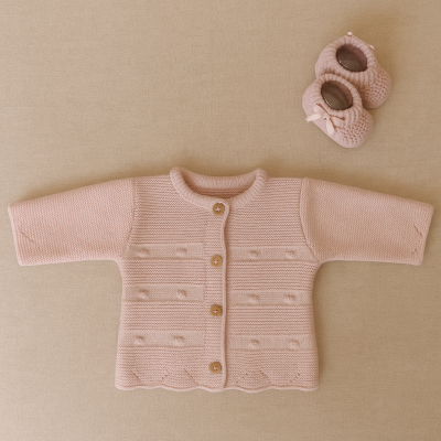Casaco de malha rosa claro para bebé com botões e botas rosa com laços sobre fundo bege