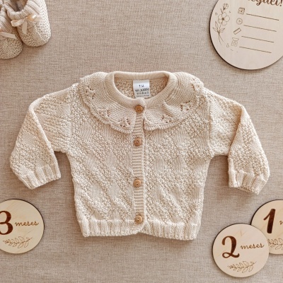 Cardigan bege tricotado para bebé com botões de madeira e discos de madeira indicativos de idade.