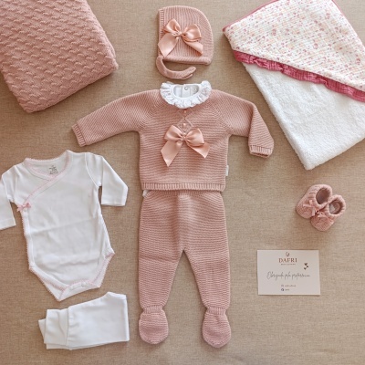 Conjunto de roupa de bebé rosa e branco com laços e manta sobre fundo bege.