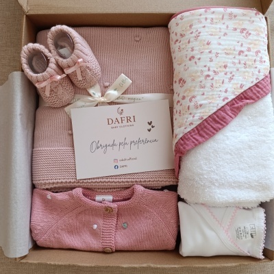 Conjunto de roupa de bebé rosa com sapatinhos de malha, camisola, manta e body dentro de uma caixa de cartão aberta