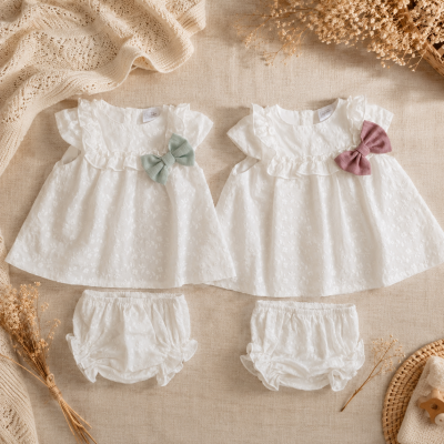 Conjuntos brancos de roupa de bebé com bordados e laços coloridos sobre tecido bege