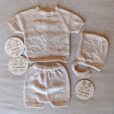 Conjunto de roupa de bebé bege composto por camisola, calções e touca em malha e discos marcadores de idade.
