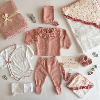 Conjunto de roupa de bebé rosa queimado e branco com manta e acessórios sobre superfície clara