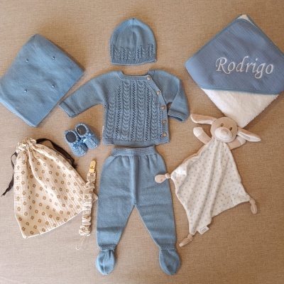 Conjunto de roupa de bebé azul com acessórios e brinquedo em fundo bege.