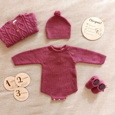 Conjunto de roupa de bebé em malha vinho com body, camisola, gorro e botinhas, e marcadores de idade em fundo bege.
