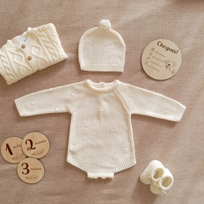 Conjunto de roupa de bebé em malha creme com acessórios e placas de madeira sobre fundo bege