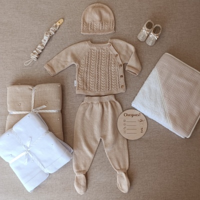 Conjunto bege de roupa de bebé em malha com gorro, cardigã e calças, sapatinhos, chupeta com fita, mantas bege e branca e placa de madeira com texto