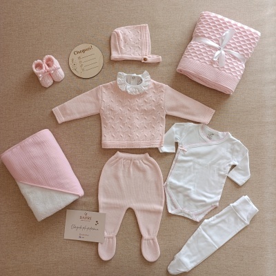 Conjunto de roupa e acessórios para bebé em tons de rosa e branco sobre superfície bege.