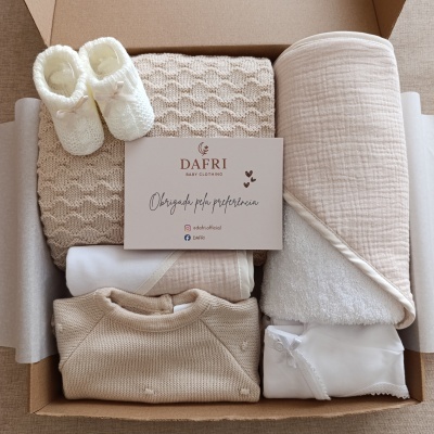 Conjunto de roupa e acessórios para bebé em tons creme e bege organizado numa caixa de cartão