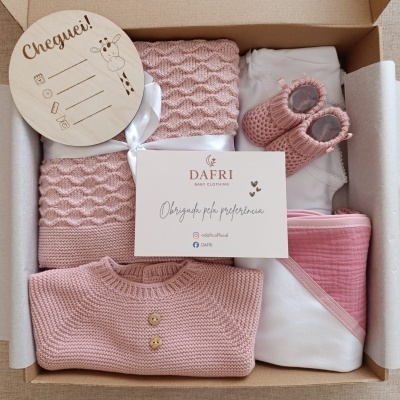Conjunto de roupa e acessórios para bebé em tons de rosa e branco dentro de caixa de presente com cartão e placa de madeira