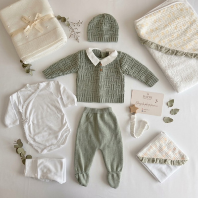 Conjunto de roupa de bebé em malha verde, body branco, cobertores e chupeta de madeira com cartão em fundo branco.
