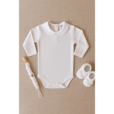 Body branco de manga comprida com gola redonda, chupeta com clipe e sapatinhos brancos sobre fundo bege.