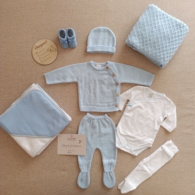 Conjunto de roupa de bebé azul-claro e branco com acessórios organizados sobre tecido bege.