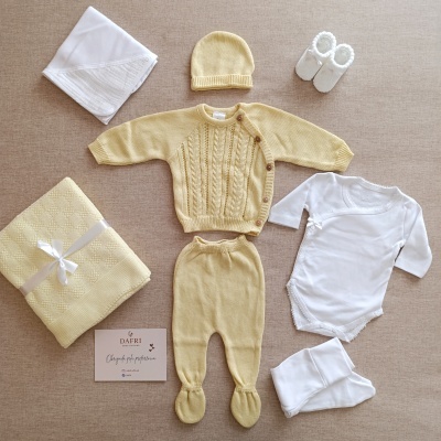 Conjunto de roupa de bebé amarelo e branco com manta e sapatinhos, dispostos em fundo bege.