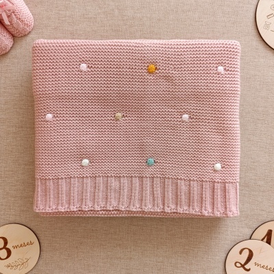 Manta rosa em malha com pompons coloridos sobre superfície bege com discos de madeira dos meses