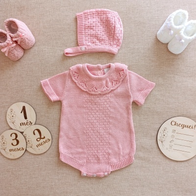 Conjunto de roupa de bebé rosa em malha com body, gorro e sapatinhos acompanhado de discos de madeira com inscrições mês e chegada.