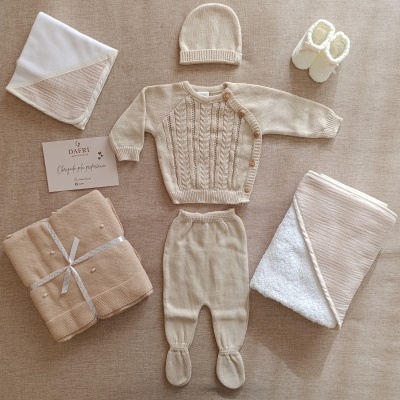 Conjunto de roupa de bebé bege de malha com casaco, calças, gorro, mantas e pantufas brancas num fundo bege.