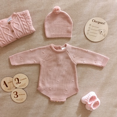 Conjunto de roupa de bebé rosa composto por body de malha, camisola com botões, gorro com pompom e sapatinhos de malha sobre tecido bege, placas de madeira para meses e placa redonda 'Cheguei!' com coala.