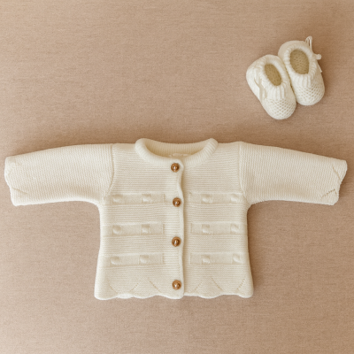 Casaco de bebé creme em tricô com botões de madeira e sapatinhos creme em tricô sobre fundo bege