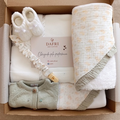 Conjunto de roupa de bebé com sapatinhos brancos, casaco verde claro, manta com padrão floral e faixa elástica, em caixa de cartão.
