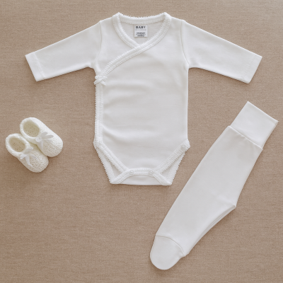 Conjunto de roupa de bebé branco com body manga comprida, leggings com pés e sapatinhos sobre fundo bege