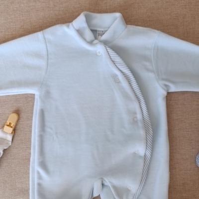 Macacão de bebé azul claro com botões e borda listrada em tecido sobre fundo bege