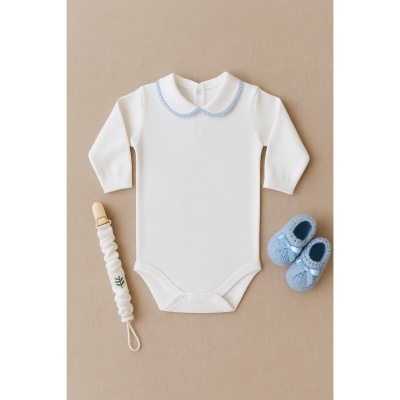 Body branco para bebé com gola azul, sapatinhos azuis e prendedor de chupeta branco sobre fundo bege