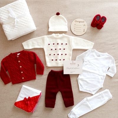 Conjunto de roupa de bebé em tons branco e vermelho, sobre fundo bege.