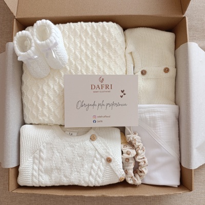 Caixa com conjunto de roupa de bebé branca em malha, sapatinhos, faixa para cabelo e manta com cartão DAFRI.