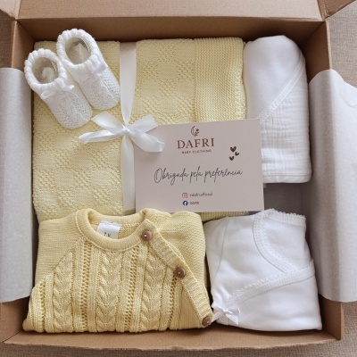 Conjunto de roupa para bebé com casaco amarelo de malha, manta amarela, sapatinhos brancos e roupa branca, numa caixa de cartão.
