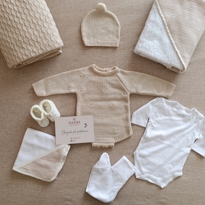Conjunto de roupa e acessórios para bebé em tons bege e branco sobre fundo bege.
