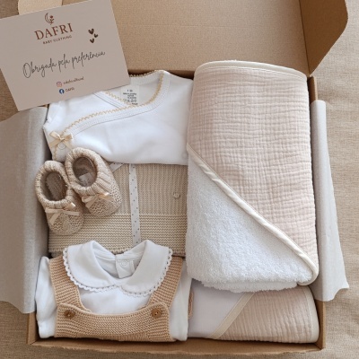 Conjunto de roupa de bebé em bege e branco com manta e sapatinhos numa caixa de cartão.