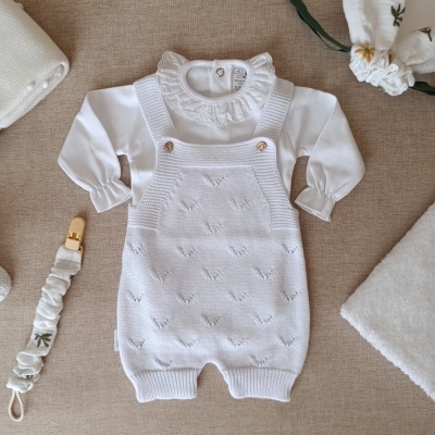 Roupa de bebé branca com jardineira de malha e camisa com gola franzida, prendedor de chupeta e pano branco floral