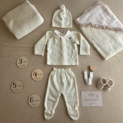 Conjunto de roupa de bebé branca com mantinha e acessórios em fundo bege.