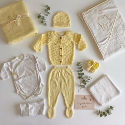 Conjunto de roupa de bebé amarelo e branco com peças em malha e algodão, manta e toalha bordada, com cartão de marca DAFRI e eucalipto decorativo.
