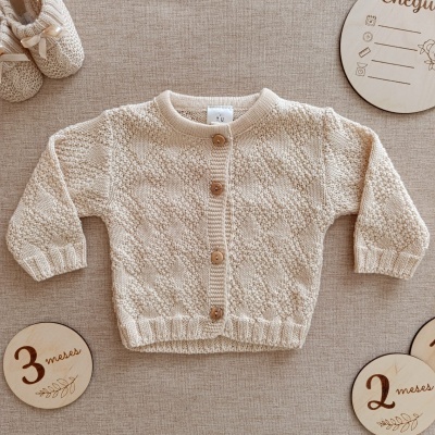 Cardigan de bebé bege em malha com botões de madeira e sapatinhos de malha, acompanhados por placas redondas de madeira com texto.