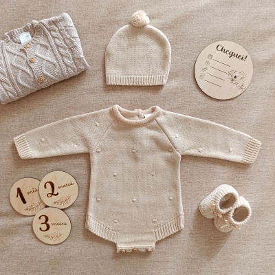 Conjunto de roupa de bebé em malha bege com body, gorro com pompom, sapatinhos em malha e acessórios de madeira