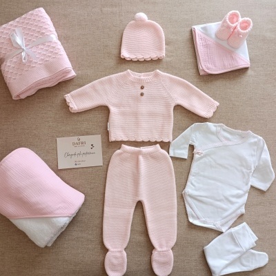 Conjunto de roupa de bebé rosa e branco com mantas e sapatinhos sobre fundo bege