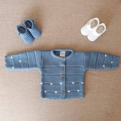 Conjunto de malha azul para bebé com casaco e dois pares de sapatinhos, num fundo bege.