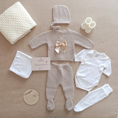 Conjunto de roupa de bebé em malha cinzenta e branco, manta branca e sapatinhos creme num fundo bege.