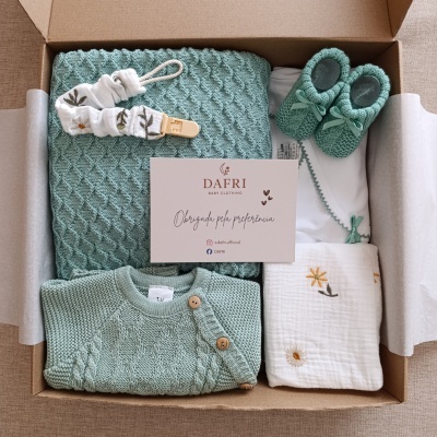 Conjunto de roupa e acessórios para bebé em tons verdes e branco com manta, casaco, sapatinhos, babete e clip para chupeta numa caixa de cartão.