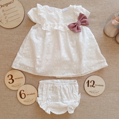 Conjunto de roupa bebé branca com vestido com folhos e laço rosa e cuecas, sobre tecido castanho, perto de discos de madeira com medidas de idade.