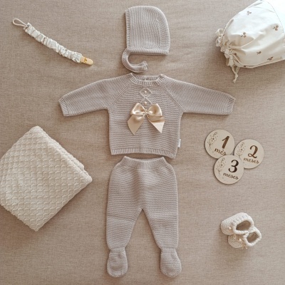 Conjunto de roupa de bebé em malha cinzenta clara com laço de cetim e acessórios em fundo bege.